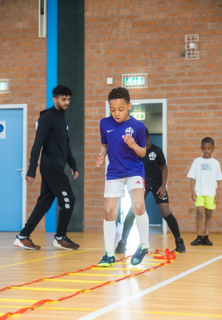 Speler van DPC Academy traint coördinatie met een loopladder onder begeleiding van een trainer.